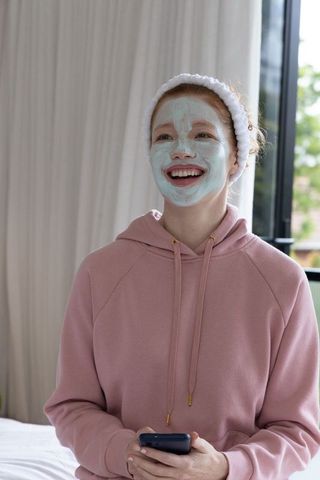 Teen woman wearing mint facial mask and pink hoodie holding smartphone smiling skincare