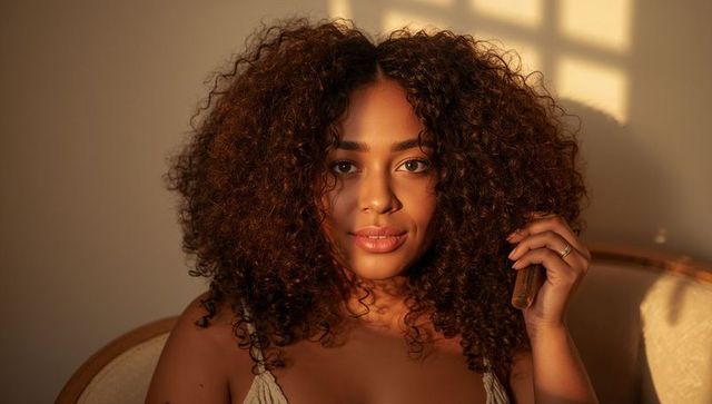 Golden-hour curly-hair portrait holding small vial near face, intimate natural beauty