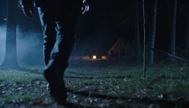 Walking toward lantern-lit canvas tent in misty night forest, lone hiker approaching