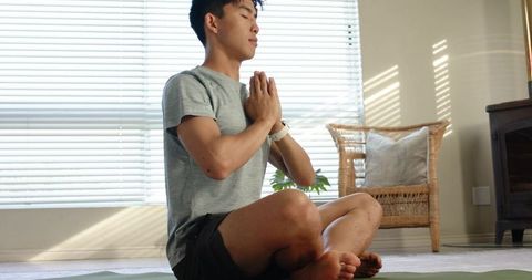 Young asian man meditating on yoga mat with smartwatch by sunlit window blinds