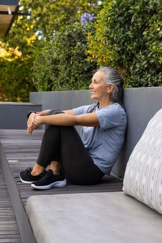 Sunlit senior woman relaxing on patio bench with smartwatch and lush garden