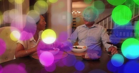 Smiling mature couple dining at home, sharing conversation over wine with bokeh lights