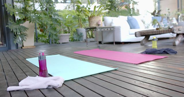 Outdoor setup perfect for yoga or fitness practices featuring pastel-colored mats, water bottles, and towels on a wooden deck. The surrounding greenery and pillows on benches create a serene and calming environment suitable for promoting relaxation and well-being imagery. Ideal for fitness blogs, wellness advertisements, environmental lifestyle content, or articles focusing on home workout routines.
