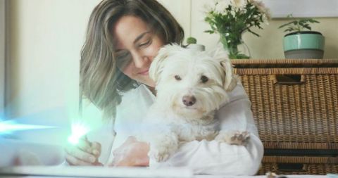 Smiling woman holding fluffy white dog at sunlit home desk, cozy pet bonding moment