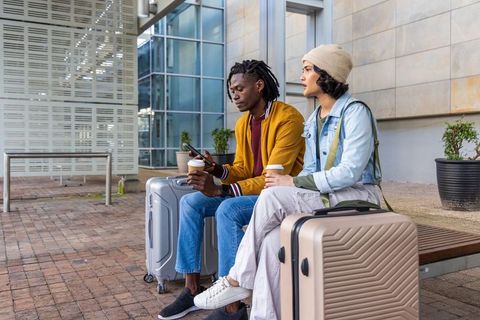 Diverse couple waiting with luggage and coffee outside transport hub, travel lifestyle