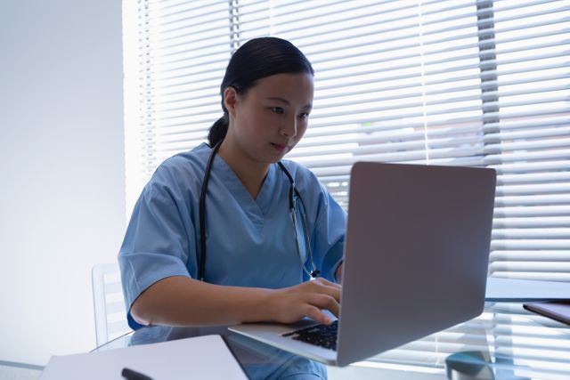 Female surgeon working on laptop at desk in hospital clinic. Ideal for illustrating modern healthcare, medical technology, and professional healthcare environments. Useful for medical websites, healthcare blogs, and educational materials.