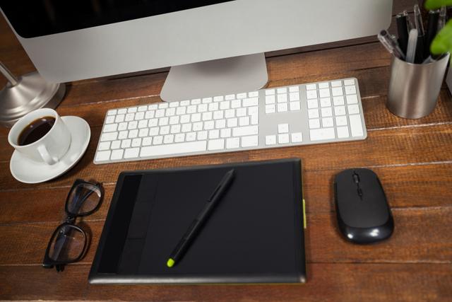 Organized office workspace featuring a desktop computer, graphic tablet with stylus, keyboard, mouse, coffee cup, glasses, and office supplies. Ideal for illustrating productivity, creative work environments, modern office setups, or home office organization tips.