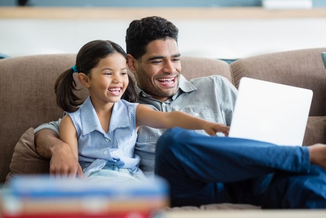 This image depicts a father and daughter sitting on a sofa, smiling and looking at a laptop. Ideal for use in articles or advertisements related to family bonding, use of technology in homes, online learning, parenting tips, or leisure time activities. It can also be used to illustrate family dynamics in blog posts, social media content, or educational materials.