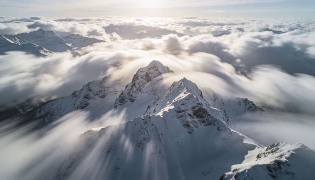 Sunlight Casting Rays Over Jagged Alpine Peaks and Drifting Clouds From Aerial Summit View