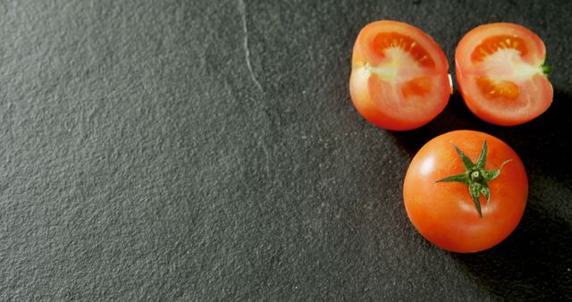 Fresh red tomatoes are halved and whole on a dark slate surface, with copy space. Their vibrant color and freshness suggest healthy eating and the importance of including vegetables in one's diet.
