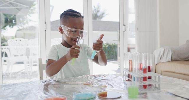 Young Black boy wearing safety goggles conducting a science experiment with various colorful liquids in test tubes at home. He appears focused and enthusiastic about his task. This image can be used in educational materials, homeschooling guides, advertisements for science kits, and content promoting STEM education among children.