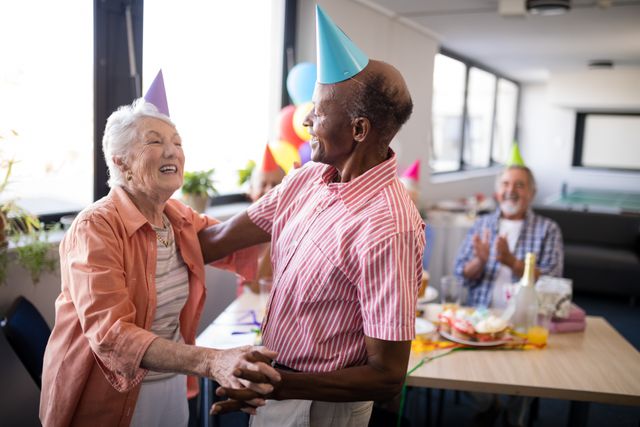 Senior couple wearing party hats dancing at birthday party in nursing home