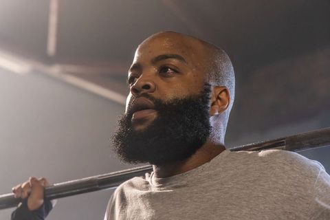 Bearded bald man lifting barbell across shoulders sweating, focusing in industrial gym