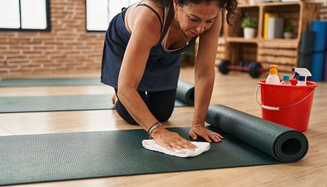 Kneeling woman cleaning yoga mat with cloth and red bucket, studio fitness hygiene routine