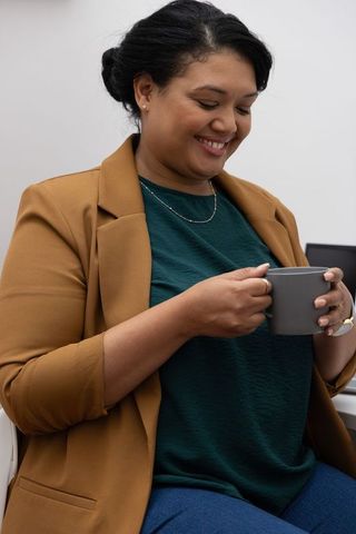 Smiling professional woman holding gray mug, relaxing at desk in mustard blazer, green top