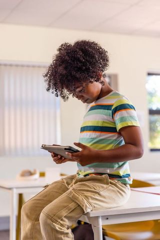 African american boy sitting on desk using tablet, focused learning in classroom