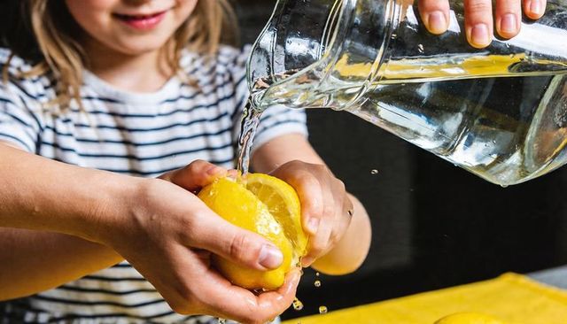 Child squeezing lemon while adult pouring water from glass pitcher, closeup fresh citrus action