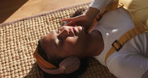 Sunlit asian woman listening to music on over-ear headphones while relaxing on woven rug