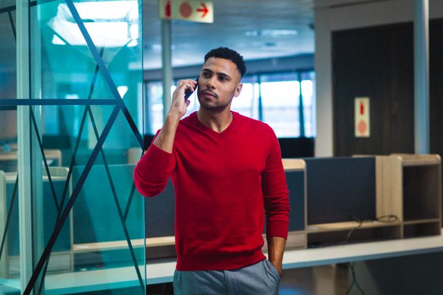 Young African American businessman standing in a modern office, answering a phone call. Ideal for use in corporate business promotions, advertisements for wireless technology, or articles about modern office environments and professional communication.