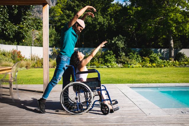 Young interracial couple enjoying a moment of dance by the poolside, with the woman in a wheelchair. This image captures love, inclusivity, and the joy of spending time together outdoors. Ideal for use in campaigns promoting diversity, disability awareness, and romantic relationships.