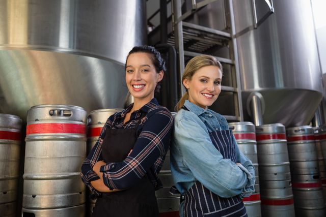 Two female coworkers standing back-to-back with arms crossed in a brewery. They are smiling and wearing work uniforms, surrounded by stainless steel tanks and beer kegs. This image can be used to represent teamwork, industrial work environments, women in brewing, and professional collaboration.