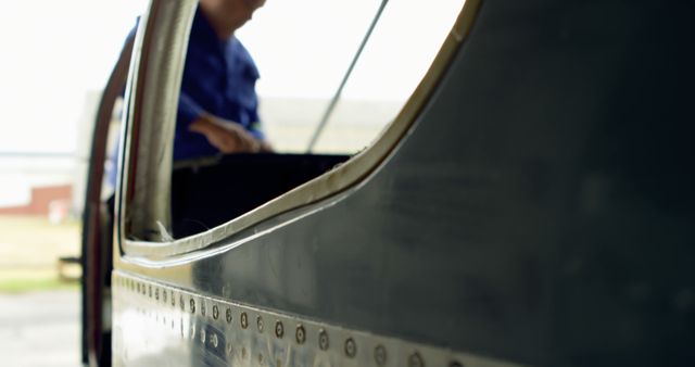 A mechanic is working on the outer structure of an aircraft in a hangar. The scene captures a close-up of the aircraft's exterior, with riveting details visible. Ideal for use in publications or advertisements relating to aviation maintenance, engineering services, aircraft repair education, and safety practices within the aviation industry.