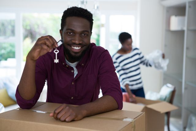Man smiling and holding key to new house while standing next to cardboard boxes. Background shows another person unpacking. Ideal for use in real estate promotions, moving services advertisements, and homeownership articles.