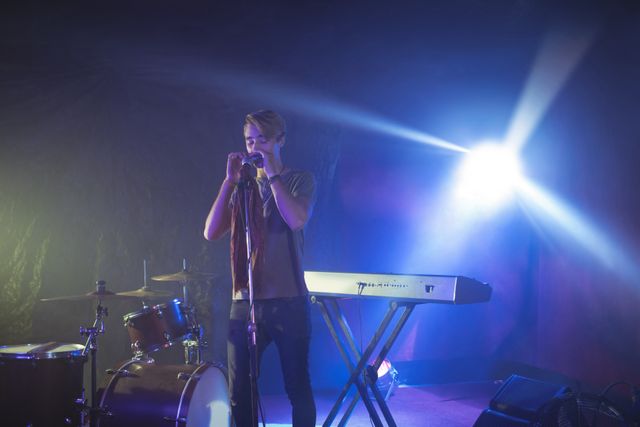 Male singer confidently performing on a brightly illuminated stage in a nightclub. He stands at a microphone with a keyboard in front of him, surrounded by stage lights and musical equipment. Ideal for use in promotions for live music events, nightclub advertisements, or articles about musicians and performances.