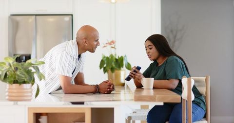 Diverse couple chatting at kitchen island, man leaning while woman showing smartphone