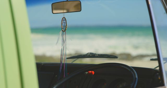 Image captures interior view of retro van with dreamcatcher hanging from rearview mirror, overlooking beach and ocean. Ideal for use in travel blogs, vacation websites, advertisements aimed at adventure-seekers or promoting van-life culture.