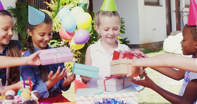 A group of diverse children is gathered at an outdoor birthday party, with one girl receiving gifts from her friends, with copy space. Their joyful expressions and party hats add to the festive atmosphere of the celebration.