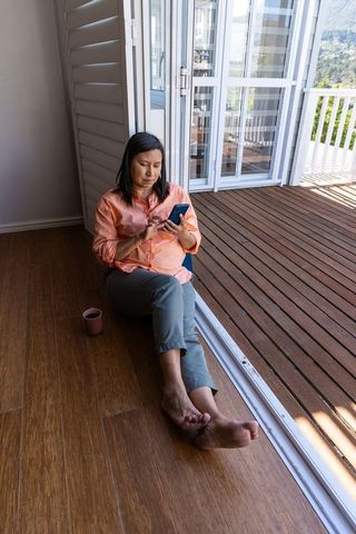 Woman relaxing barefoot on wooden floor at open doorway using smartphone with coffee mug