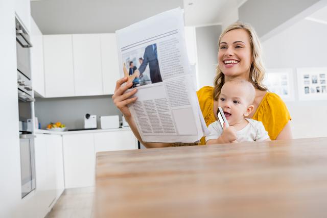Mother reading newspaper while holding baby boy in modern kitchen. Ideal for use in articles or advertisements related to parenting, family life, morning routines, or home living. Can be used in blogs, magazines, or social media posts focusing on motherhood, family bonding, or domestic happiness.