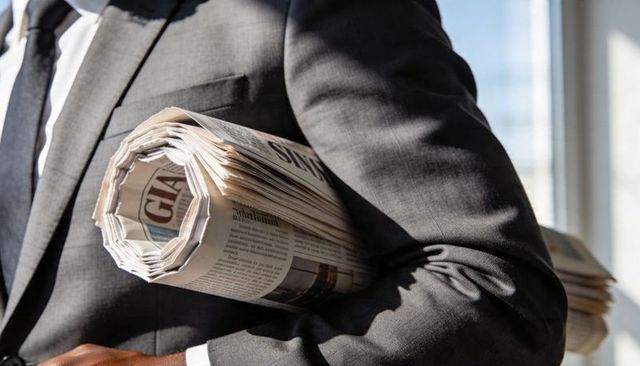 Businessman carrying rolled broadsheet newspaper under arm near sunlit office window
