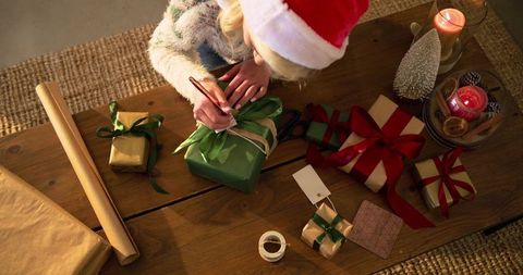 Woman writing gift tags and tying ribbons on wooden table wearing santa hat