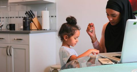 Mother in Hijab Feeding Daughter at Home Kitchen Table