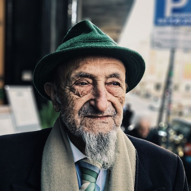 Elderly man with green hat and scarf outdoors in an urban environment, smiling slightly into the distance, conveying wisdom and dignity. Ideal for use in materials relating to elderly care, senior lifestyles, and fashion. Can be used for brochures, websites, and advertisements promoting mental wellness and aging gracefully.