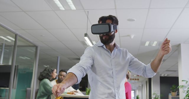 A young professional using a VR headset in a modern office. He is engaged and standing, interacting with the virtual environment. Colleagues in the background are sitting and discussing work. This image can be used to highlight technological innovation and interactive tools in contemporary business settings.