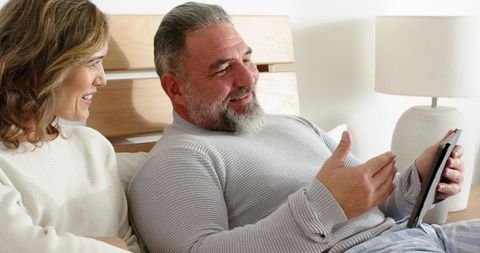 Mature couple reclining on bed sharing tablet, smiling and relaxing in cozy bedroom