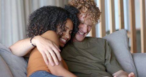 Smiling diverse couple cuddling on light sofa, sharing warm intimate moment with beaded bracelet