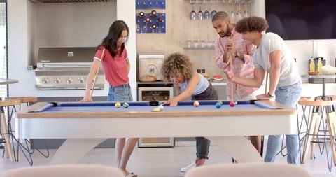 A diverse group of friends enjoys a lively game of pool in a modern indoor setting. This playful and cheerful image captures moments of friendship, social bonding, and leisure activities. Ideal for use in lifestyle blogs, social media marketing, and advertisements that highlight fun and interaction among friends.