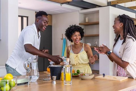 African american friends laughing, pouring drinks and preparing food at kitchen island