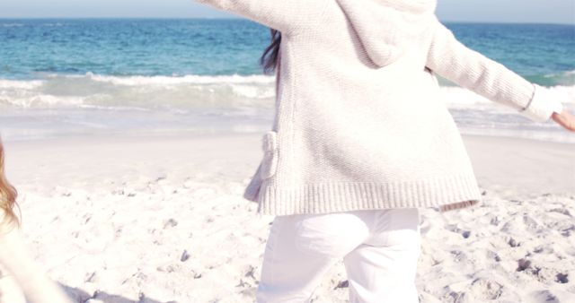 Person is joyfully walking on a sandy beach near the ocean, dressed in a cozy sweater and white pants. This image radiates relaxation and leisure, making it suitable for website banners, travel blogs, or social media posts about beach holidays, casual fashion, and outdoor activities.