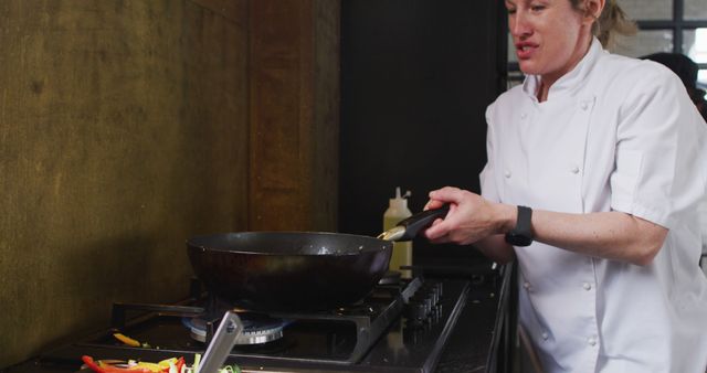 Professional chef is sautéing vegetables in a well-equipped kitchen. The person is wearing a white chef's jacket, and you can see mixed vegetables on the stove. Ideal for use in content related to culinary arts, cooking tutorials, restaurant promotion, and professional chef profiles.