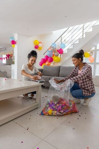 Diverse friends cleaning up after party sweeping confetti among colorful balloons