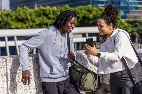 Diverse friends sharing smartphone and laughing while leaning on urban railing