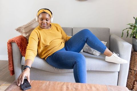 African american woman relaxing on couch listening to music in mustard sweater and headphones