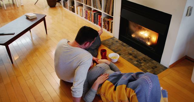 A couple enjoys a quiet moment by a warm fireplace in a modern living room. One person appears to be reading a book, while the other lies relaxed, covered with a colorful blanket. Use for concepts of relaxation, togetherness, and home comfort.