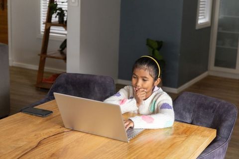 Indian girl studying on laptop at dining table, remote learning and homework concentration