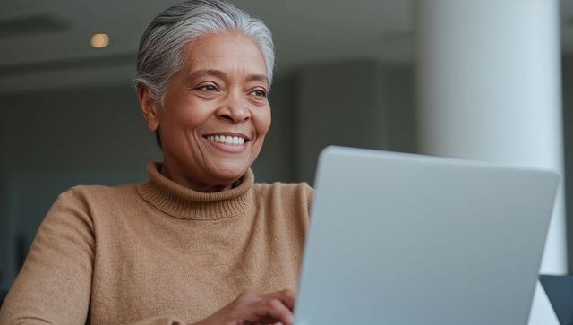 Senior woman smiling while typing on laptop in bright modern workspace brown turtleneck